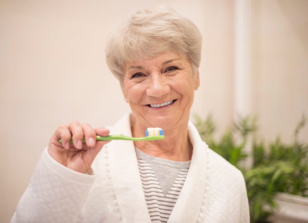 Mulher idosa sorridente segura escova de dentes verde com pasta, cuidando da vida útil de implantes na Clínica Aliança em Itabaiana.